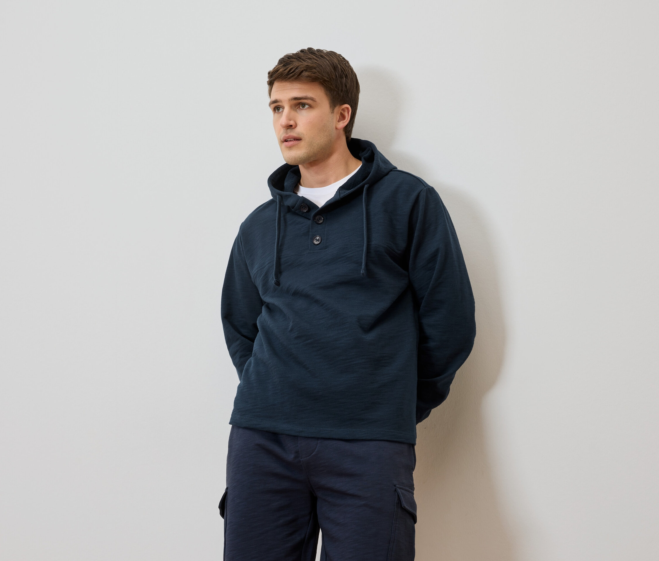 Mężczyzna w granatowej bluzie z kapturem i szortach na tle białej ściany.