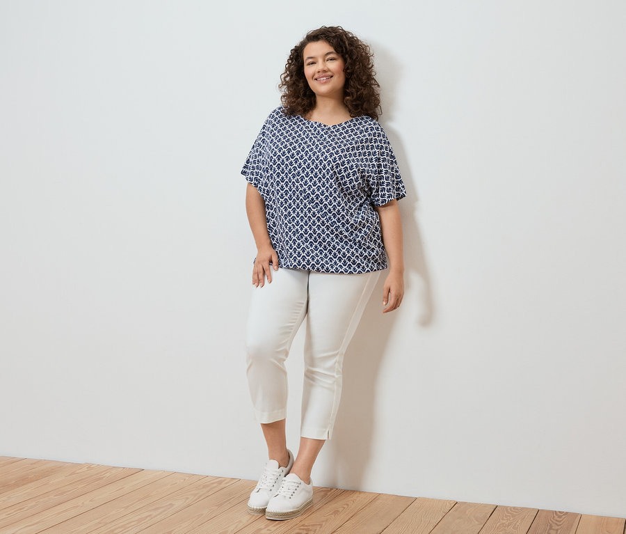 Kobieta z kręconymi włosami w bluzce w niebiesko-biały wzór, białych spodniach i białych butach sportowych stojąca na tle białej ściany.