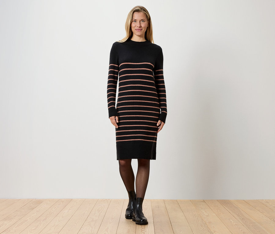 Blondynka stoi na tle białej ściany, ubrana w czarną sukienkę w paski, czarne rajstopy i czarne buty.