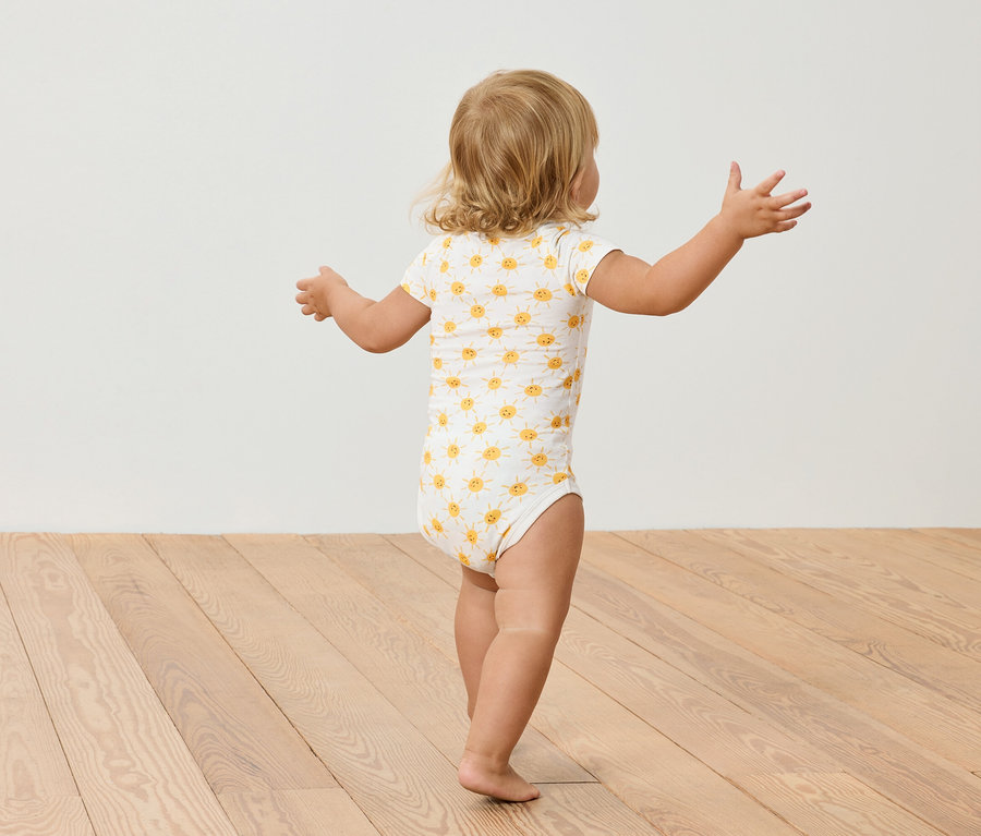 Małe dziecko z blond włosami w body w słoneczniki stoi na drewnianej podłodze.