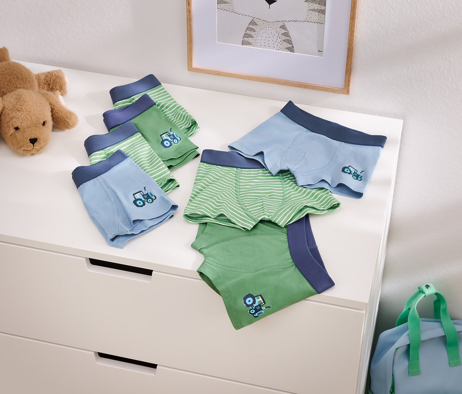 Zdjęcie przedstawia jasnoniebieskie i zielone bokserki chłopięce z motywem traktora na komodzie, obok pluszowego psa i niebieskiej torby podróżnej.