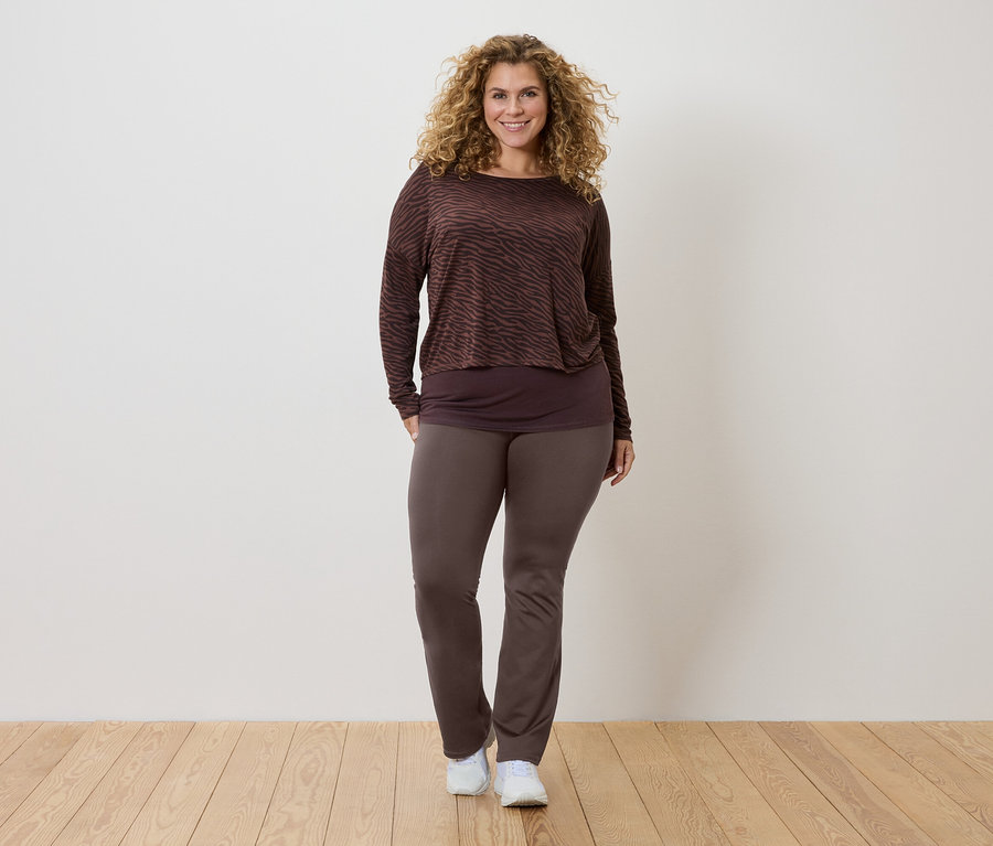 Kobieta z kręconymi włosami ma na sobie brązową bluzkę z długim rękawem we wzory, ciemniejszy top i brązowe spodnie oraz białe tenisówki.