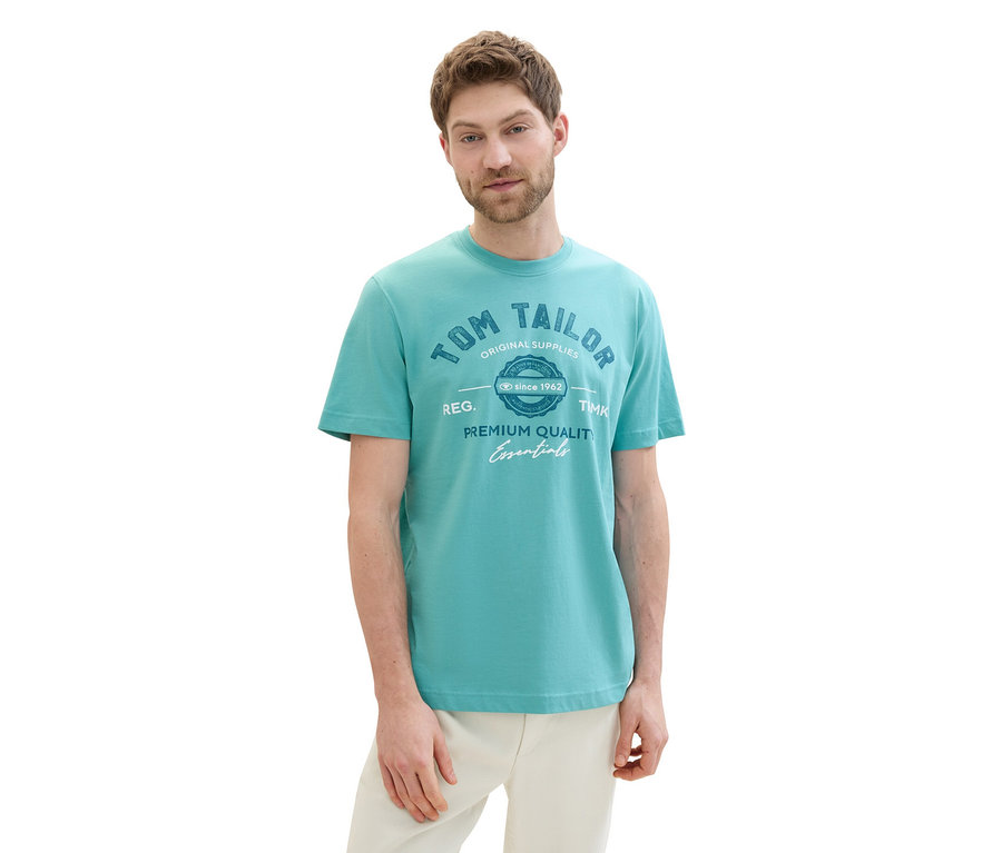 Mężczyzna ma na sobie koszulkę z logo Tom Tailor, meadow teal i jasne spodnie.