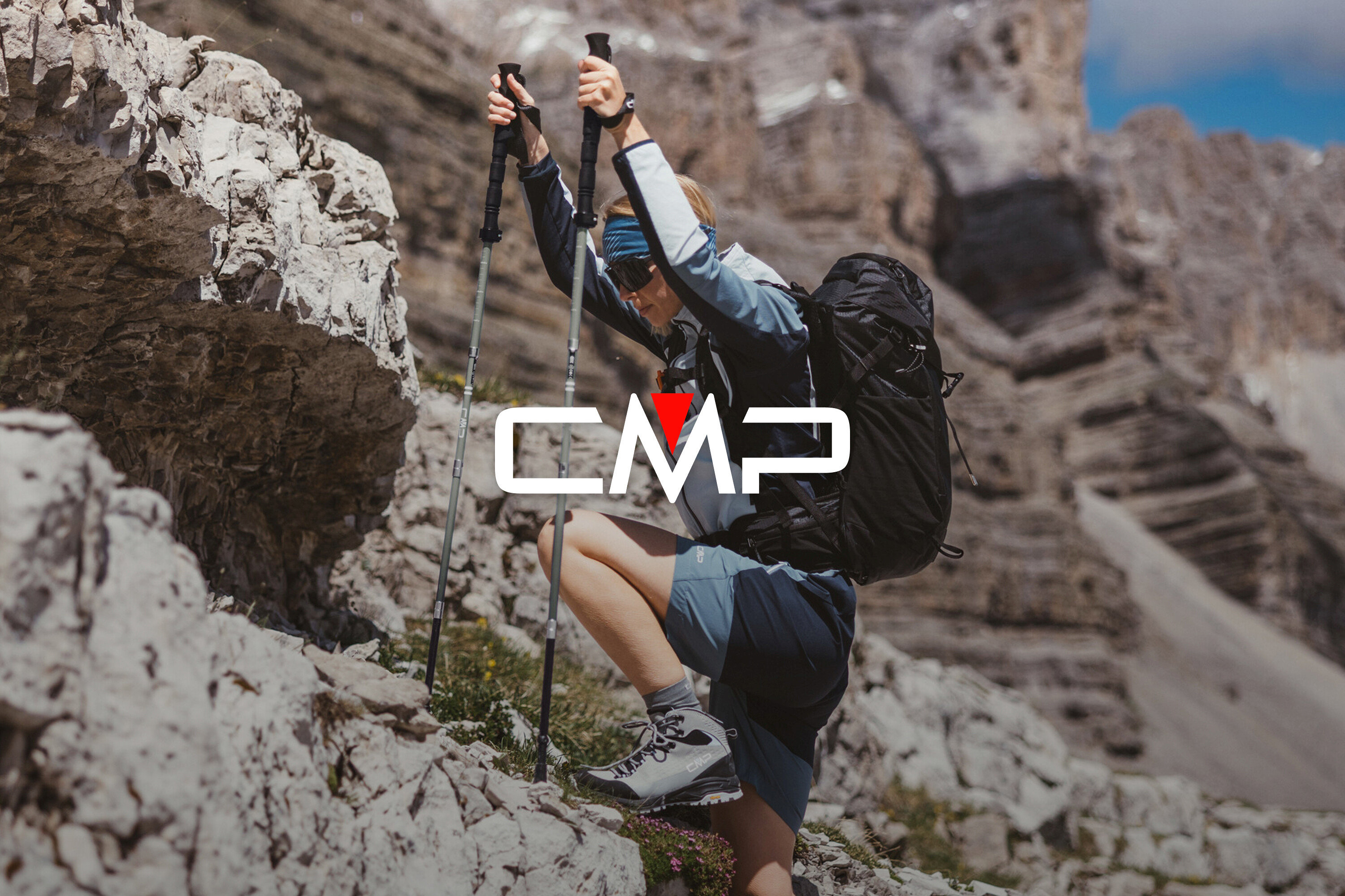 Kobieta z plecakiem i kijkami trekkingowymi wspina się po skałach. Ma na sobie buty trekkingowe, szorty i kurtkę.