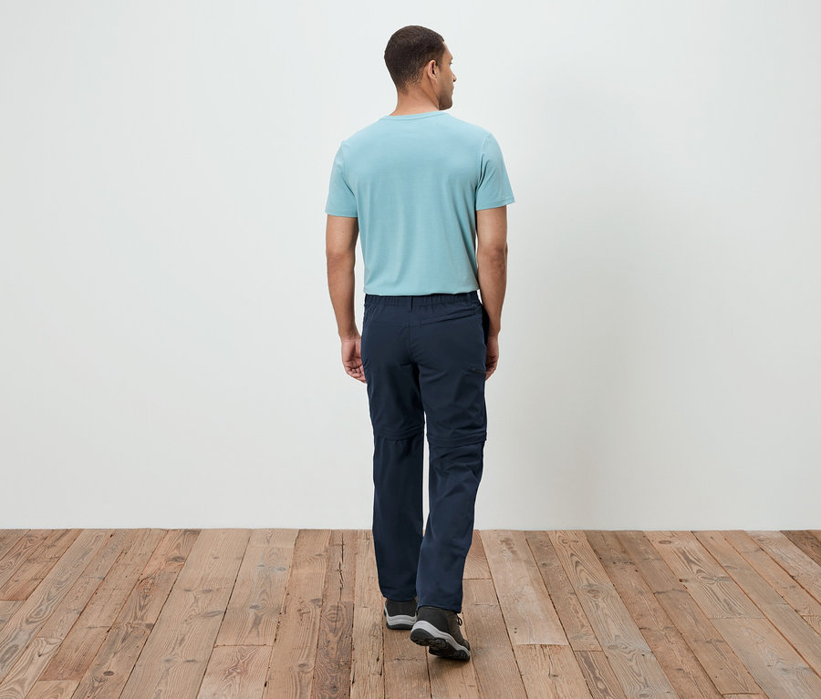Mężczyzna od tyłu, ubrany w turkusowy T-shirt i granatowe spodnie, idący po drewnianej podłodze.