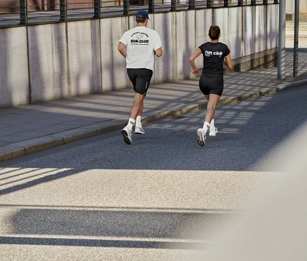 Mężczyzna w białej koszulce sportowej z napisem „RUN CLUB”, czarnych szortach sportowych i skarpetach tenisowych Coffee Crew unisex oraz kobieta w czarnej koszulce sportowej z napisem „RUN CLUB” i krótkich legginsach sportowych ActivePower biegną ulicą.