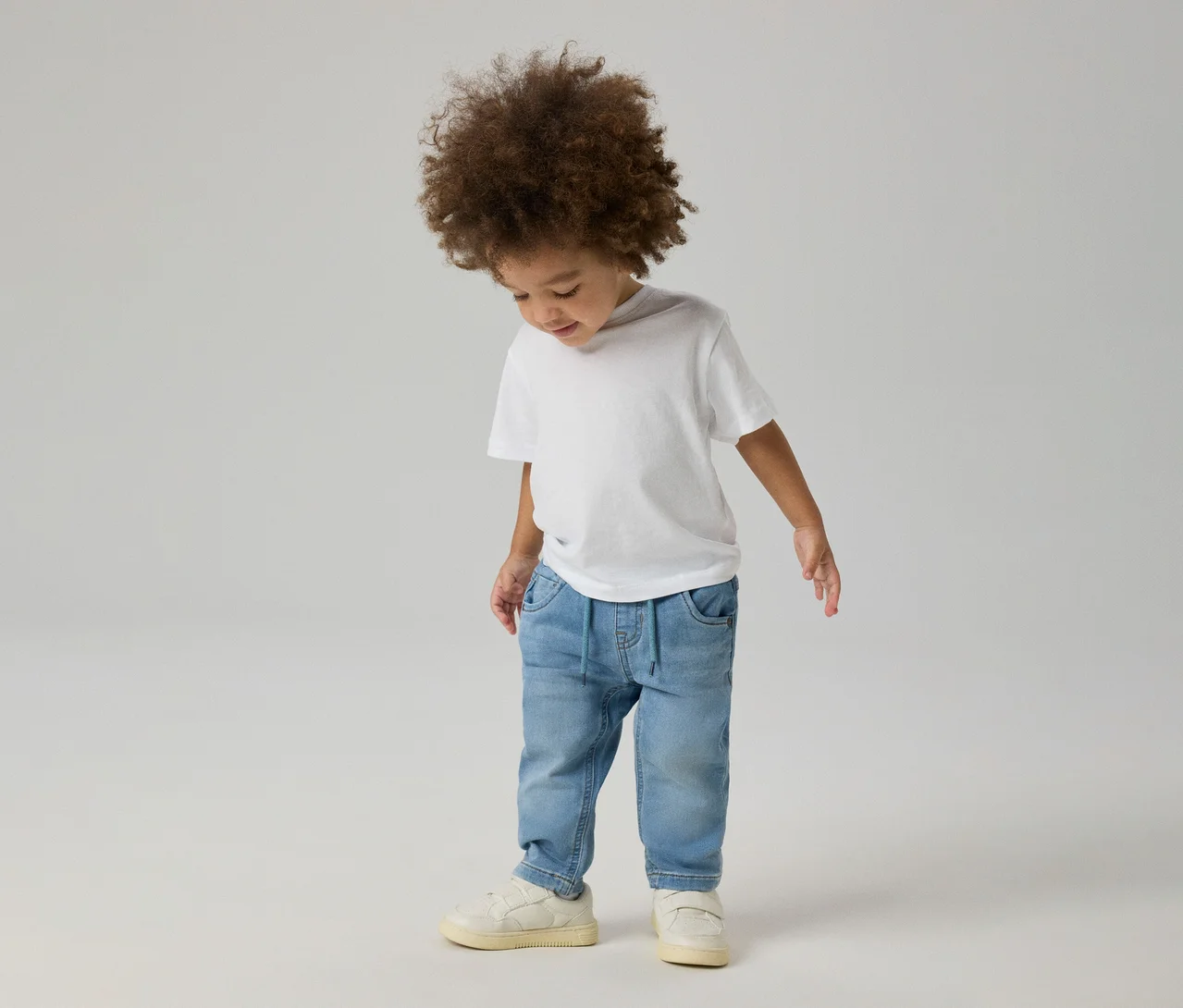 Małe dziecko z kręconymi włosami w białym T-shircie, jasnoniebieskich dżinsach NAME IT Slim Sweat Jeans i jasnych sneakersach, spoglądające w dół.
