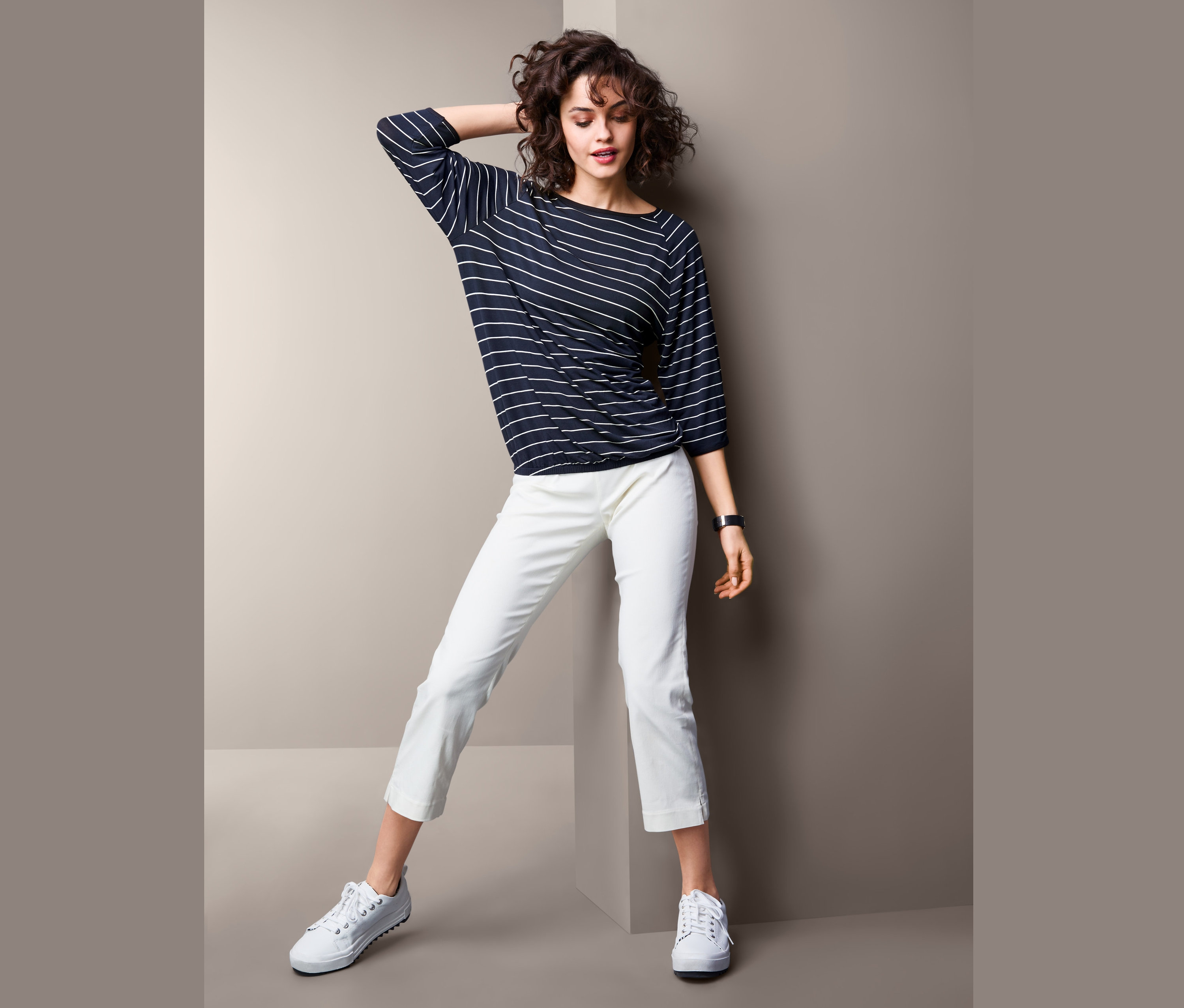 Kobieta pozuje w bluzce z kimonowymi rękawami, białych spodniach i butach sportowych.