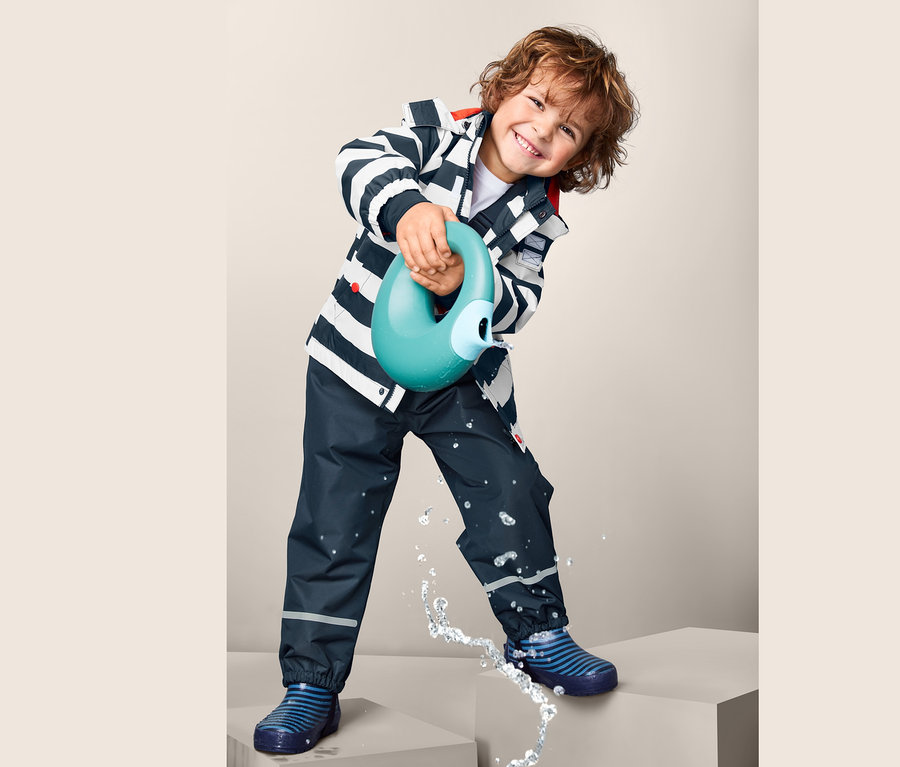 Chłopiec trzyma niebieską konewkę i leje z niej wodę. Ma na sobie spodnie przeciwdeszczowe z materiału z recyklingu i kurtkę w niebiesko-białe paski.