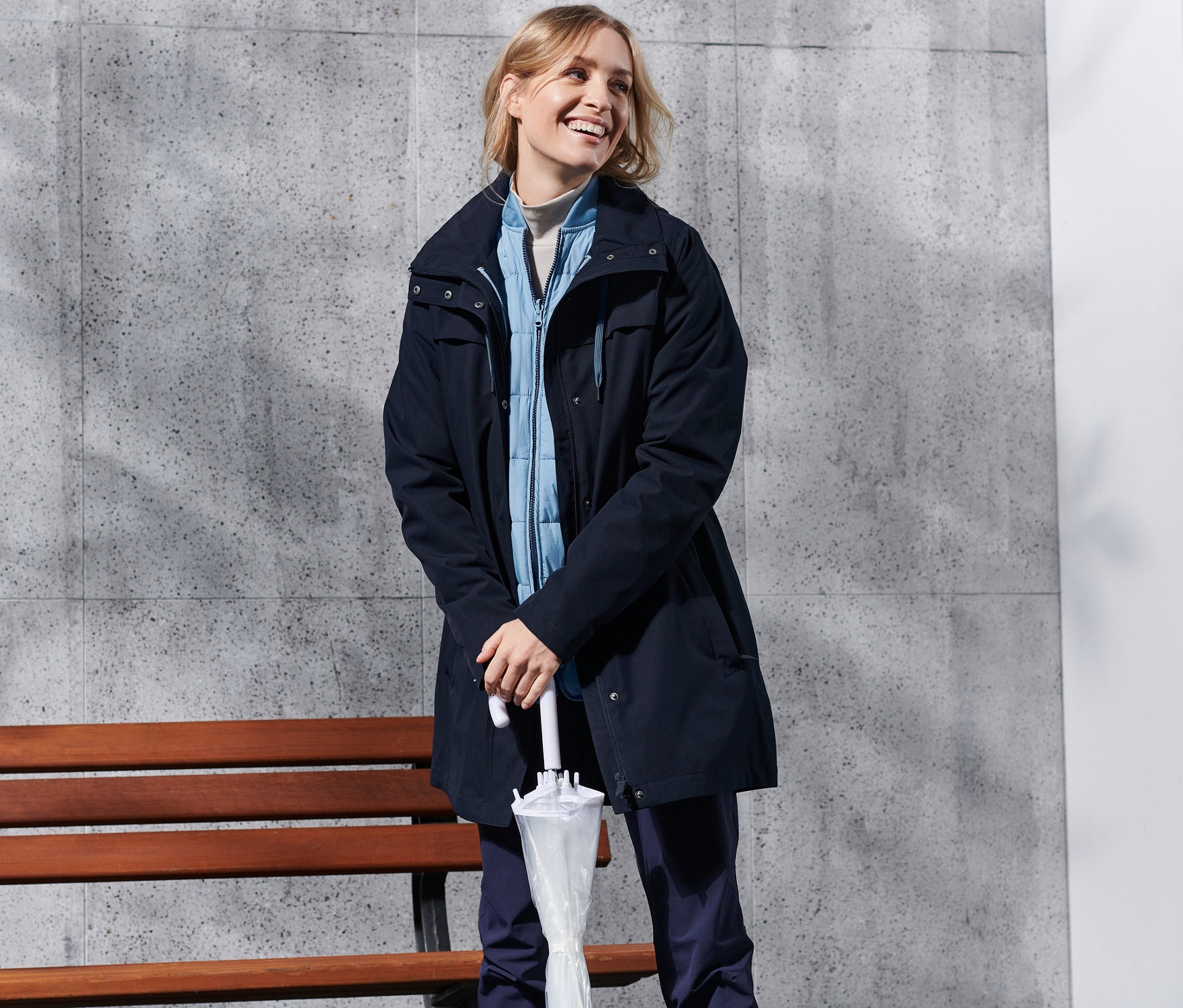 Uśmiechnięta kobieta stoi z parasolem, ubrana w niebieski płaszcz trekkingowy 3 w 1.