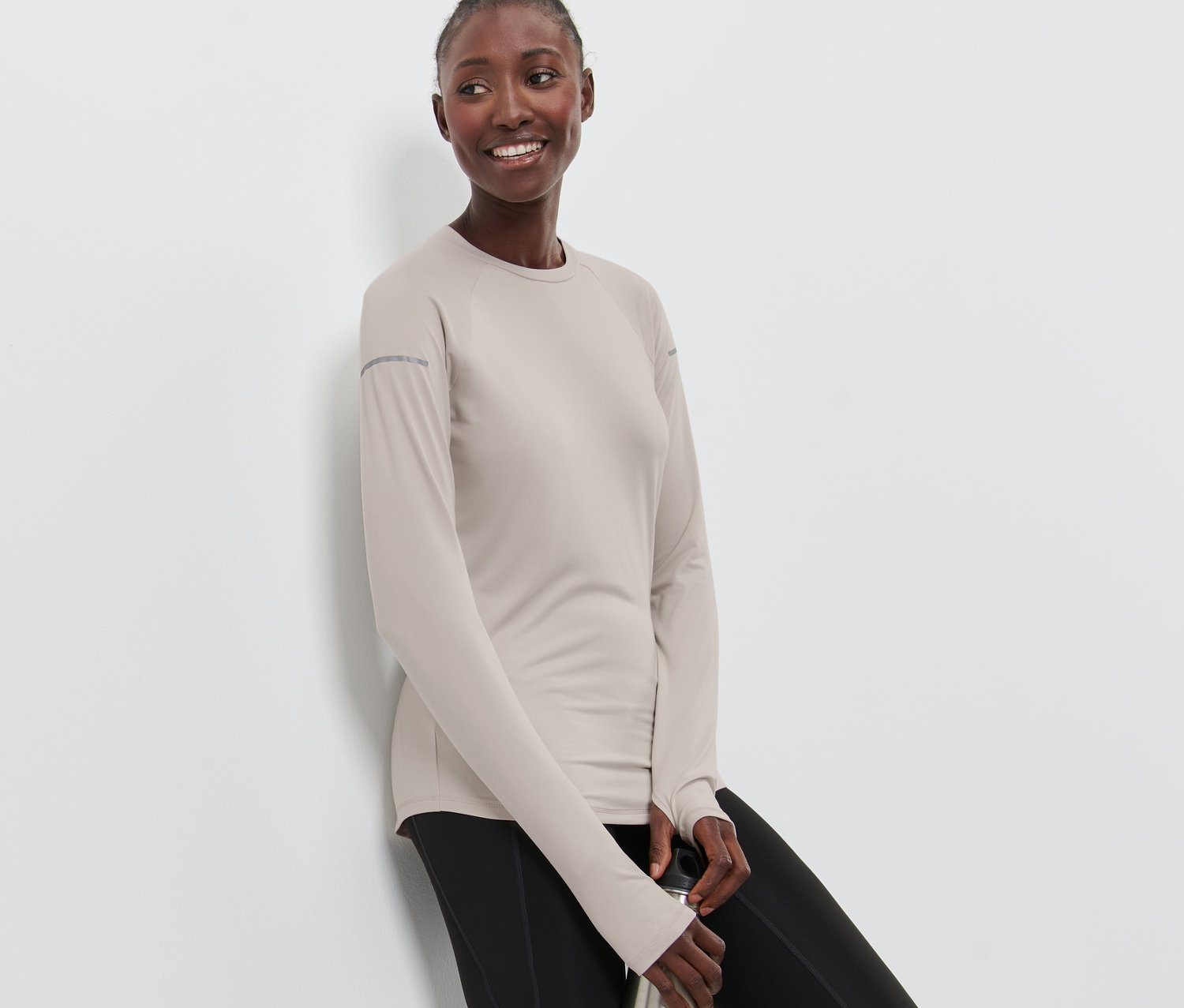 Kobieta opiera się o ścianę i trzyma metalową butelkę na wodę. Ma na sobie szarą bluzę funkcyjną z długim rękawem i czarne legginsy.