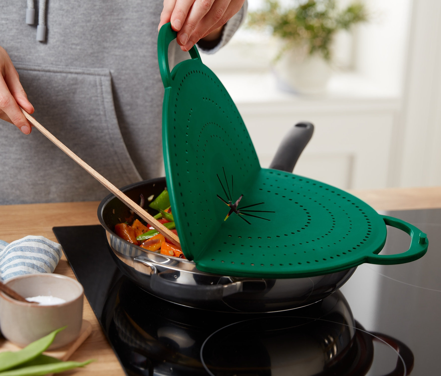 Osoba trzyma zieloną osłonę na patelnię 4 w 1 nad patelnią z jedzeniem.