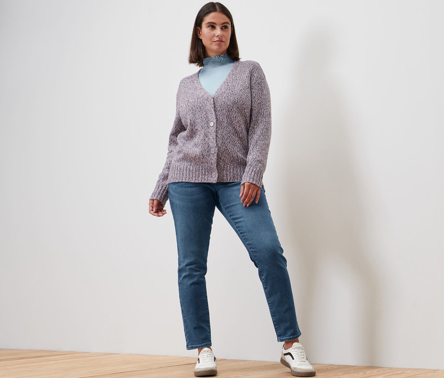Kobieta pozuje w bluzce ze stójką, szaroniebieskiej, jeansach slim »Fit Emma«, mid blue denim, rozpinanym swetrze z zawartością wełny i skórzanych sneakersach.
