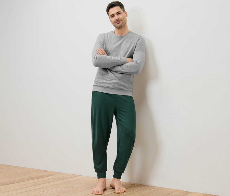 Mężczyzna pozuje ze skrzyżowanymi ramionami, ubrany w szarą piżamę i zielone spodnie od piżamy.