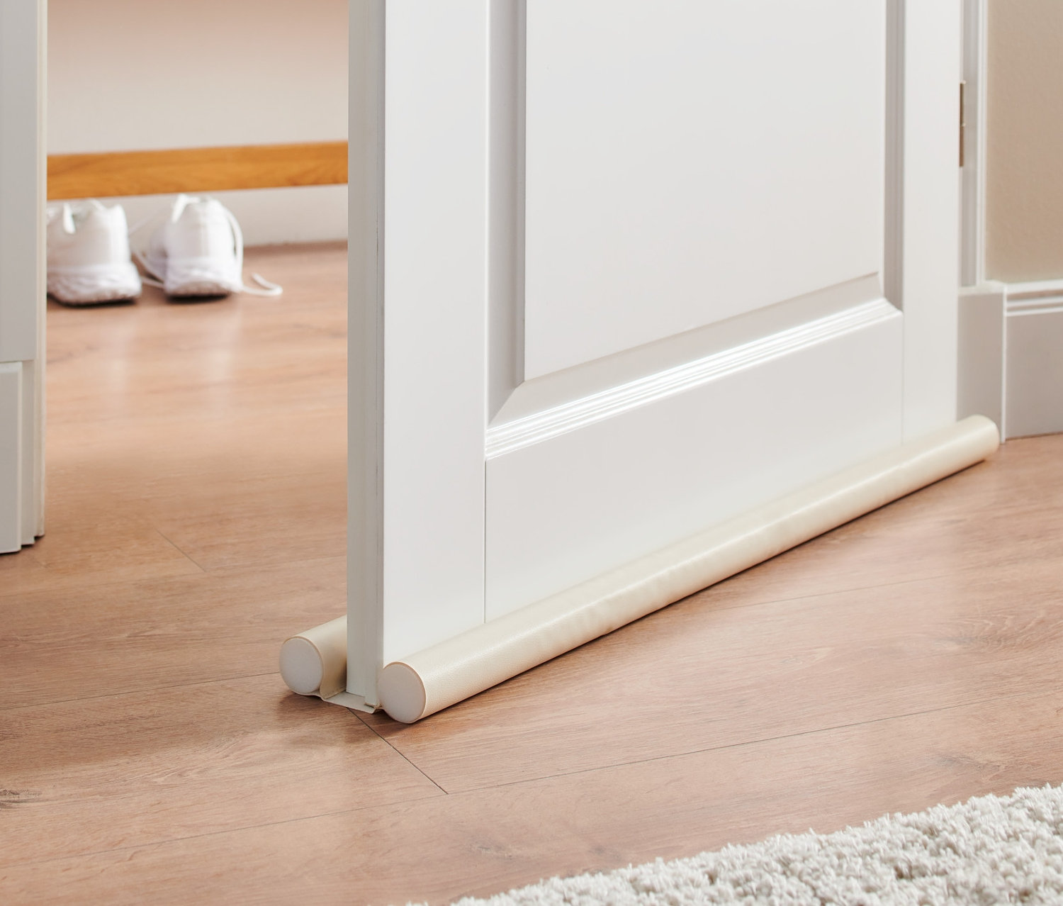 Białe drzwi z podwójną uszczelką pod drzwi na drewnianej podłodze i białym dywanie.