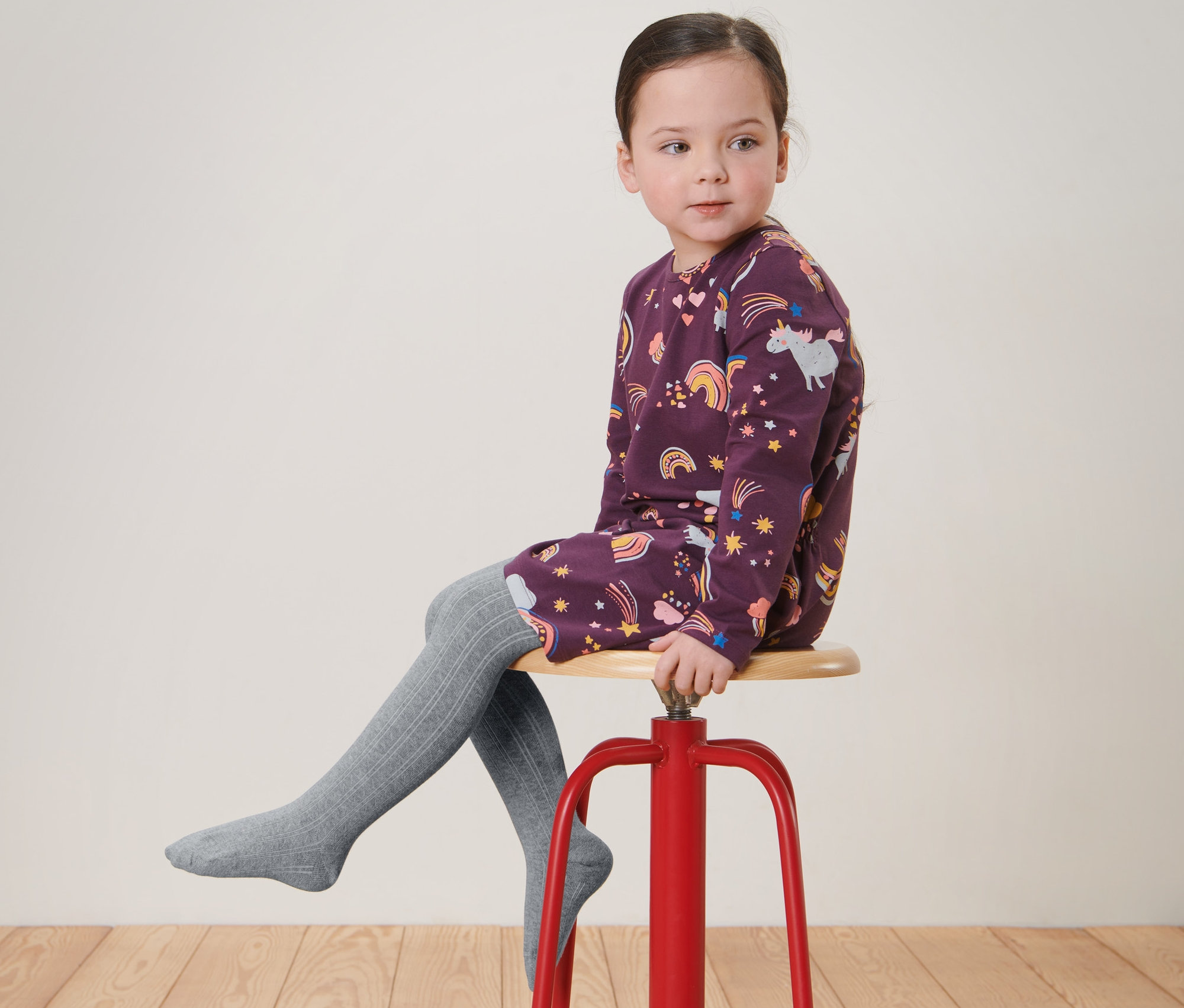 Dziewczynka siedzi na czerwonym stołku, ma na sobie sukienkę we wzory i szare Rajstopy dziecięce, 2 pary.