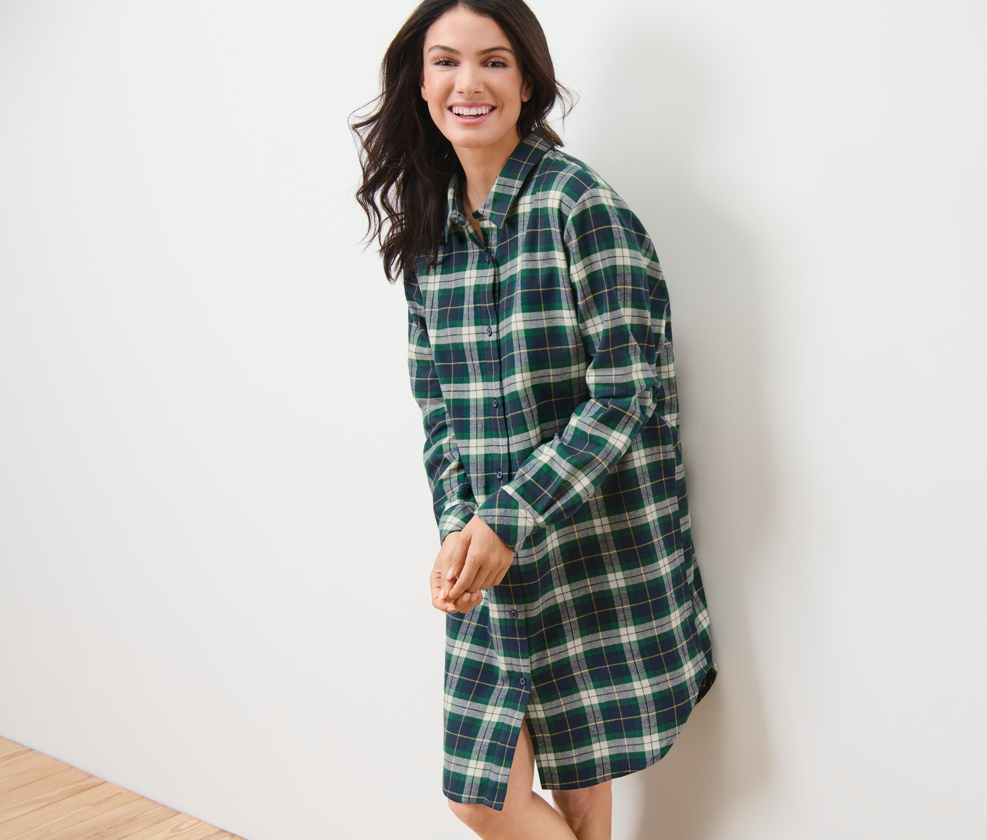 Uśmiechnięta kobieta oparta o białą ścianę ma na sobie zieloną flanelową koszulę nocną.