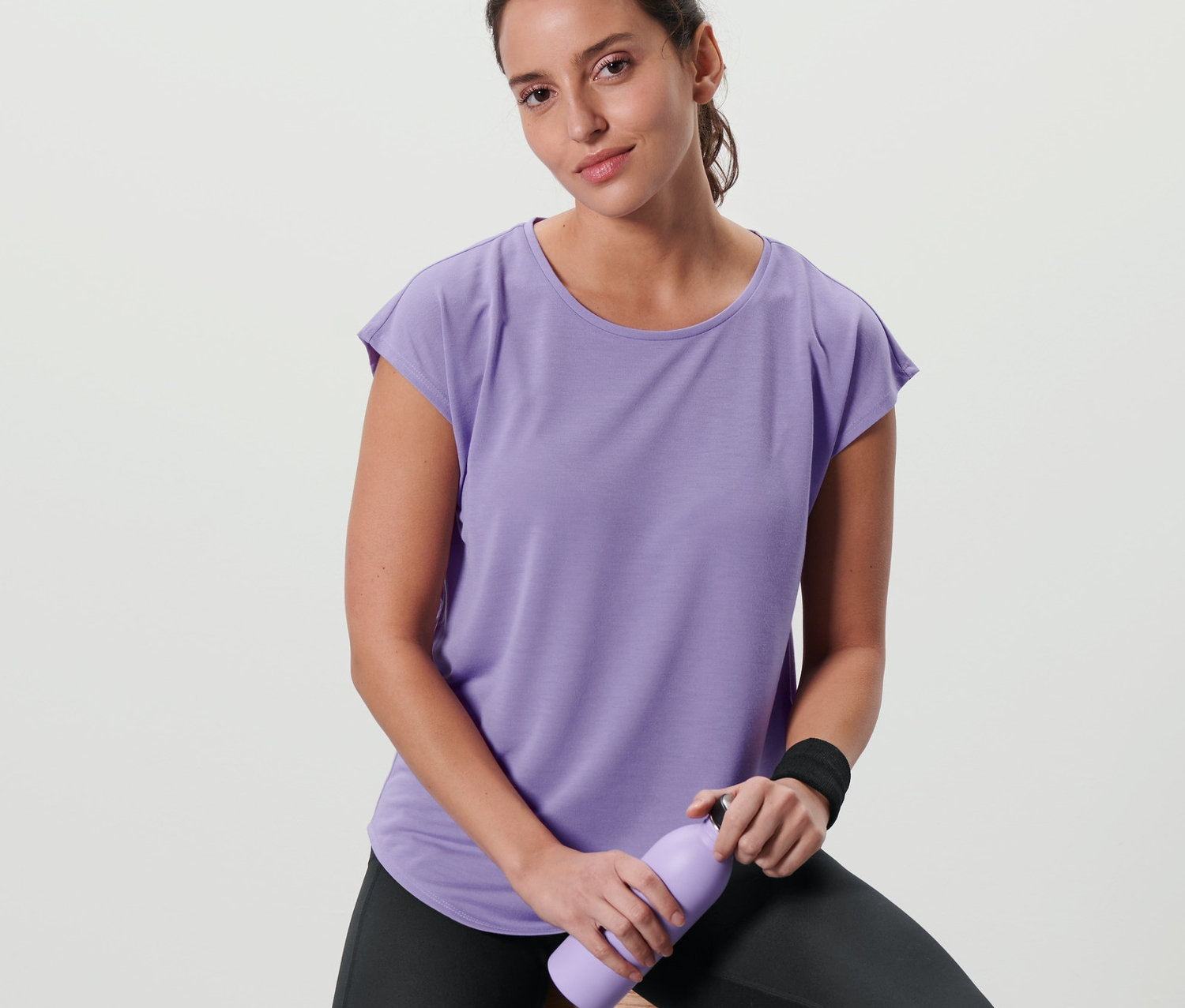 Kobieta siedzi na stołku, ubrana w liliową koszulkę sportową i trzyma fioletową butelkę.