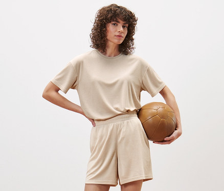 Kobieta pozuje w studio, ubrana w szorty sportowe z tkaniny frotte i trzyma brązową piłkę do piłki nożnej.