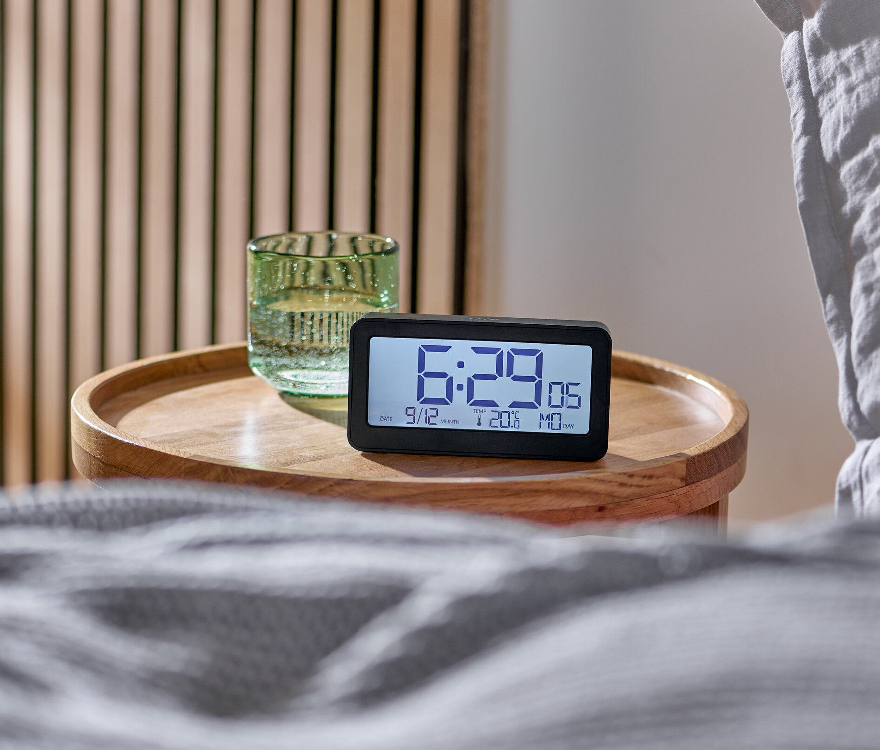 Budzik cyfrowy z wyświetlaczem LCD sterowany sygnałem radiowym stoi na drewnianym stoliku nocnym obok łóżka.