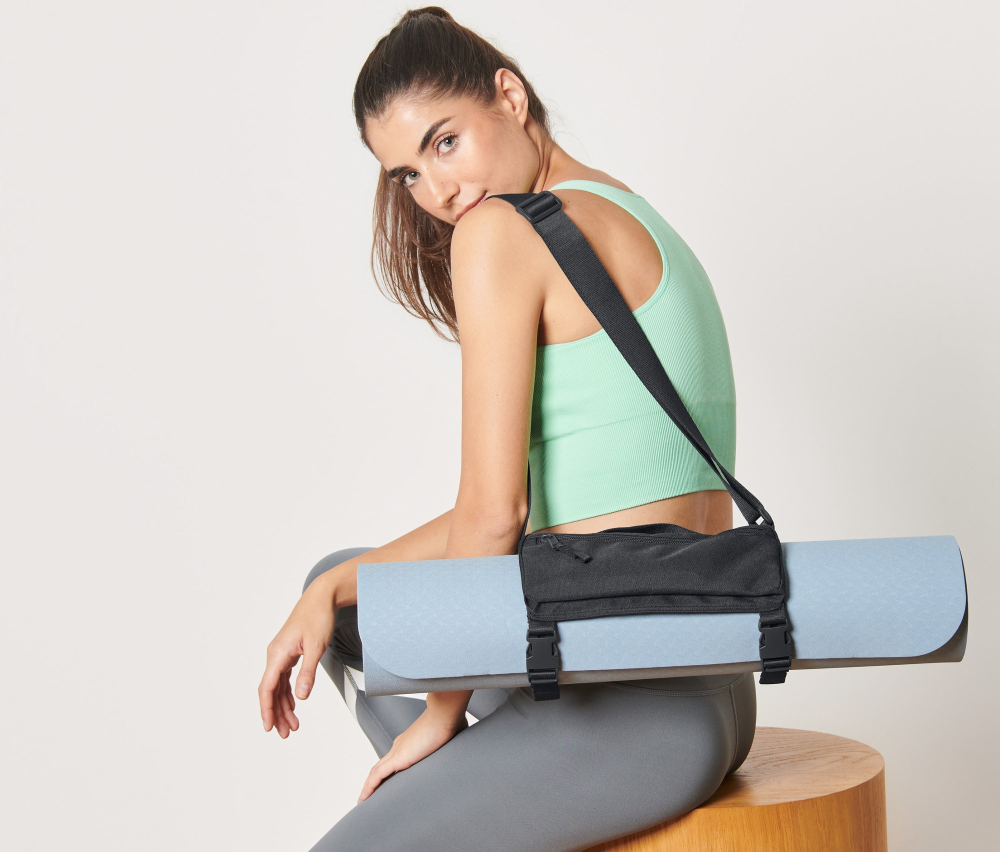 Kobieta siedzi z paskiem do noszenia maty na ramieniu, który trzyma niebieską matę do jogi.