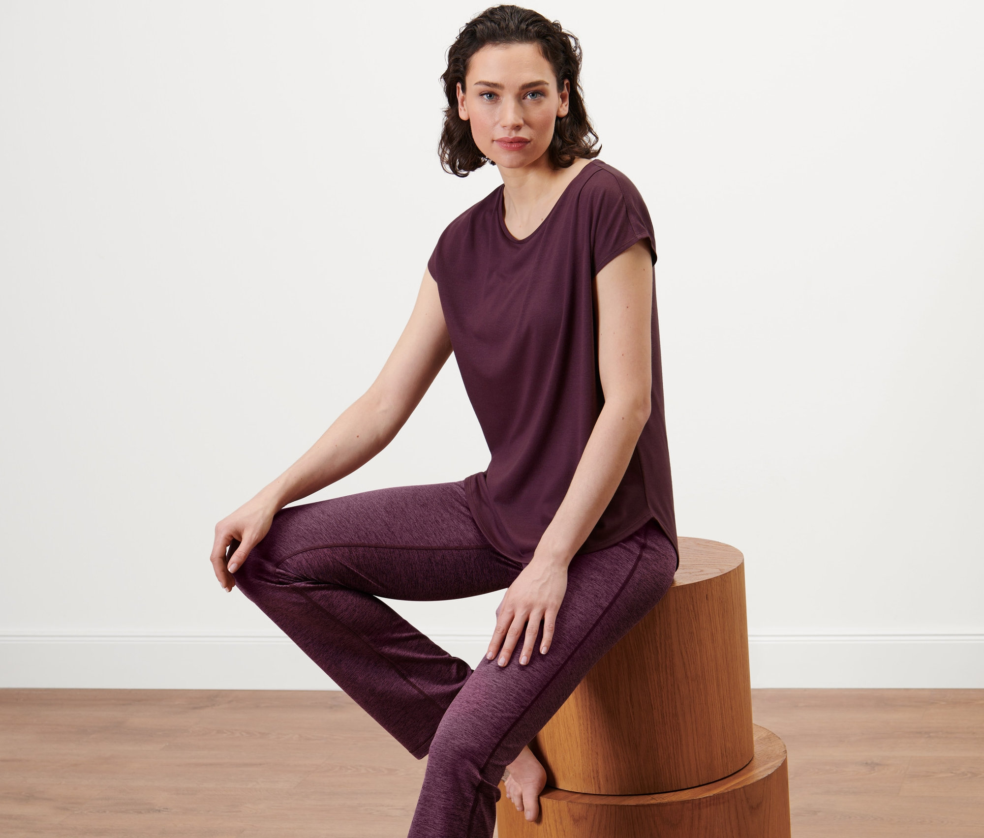 Kobieta siedzi na drewnianym stołku, ubrana w śliwkową koszulkę sportową.