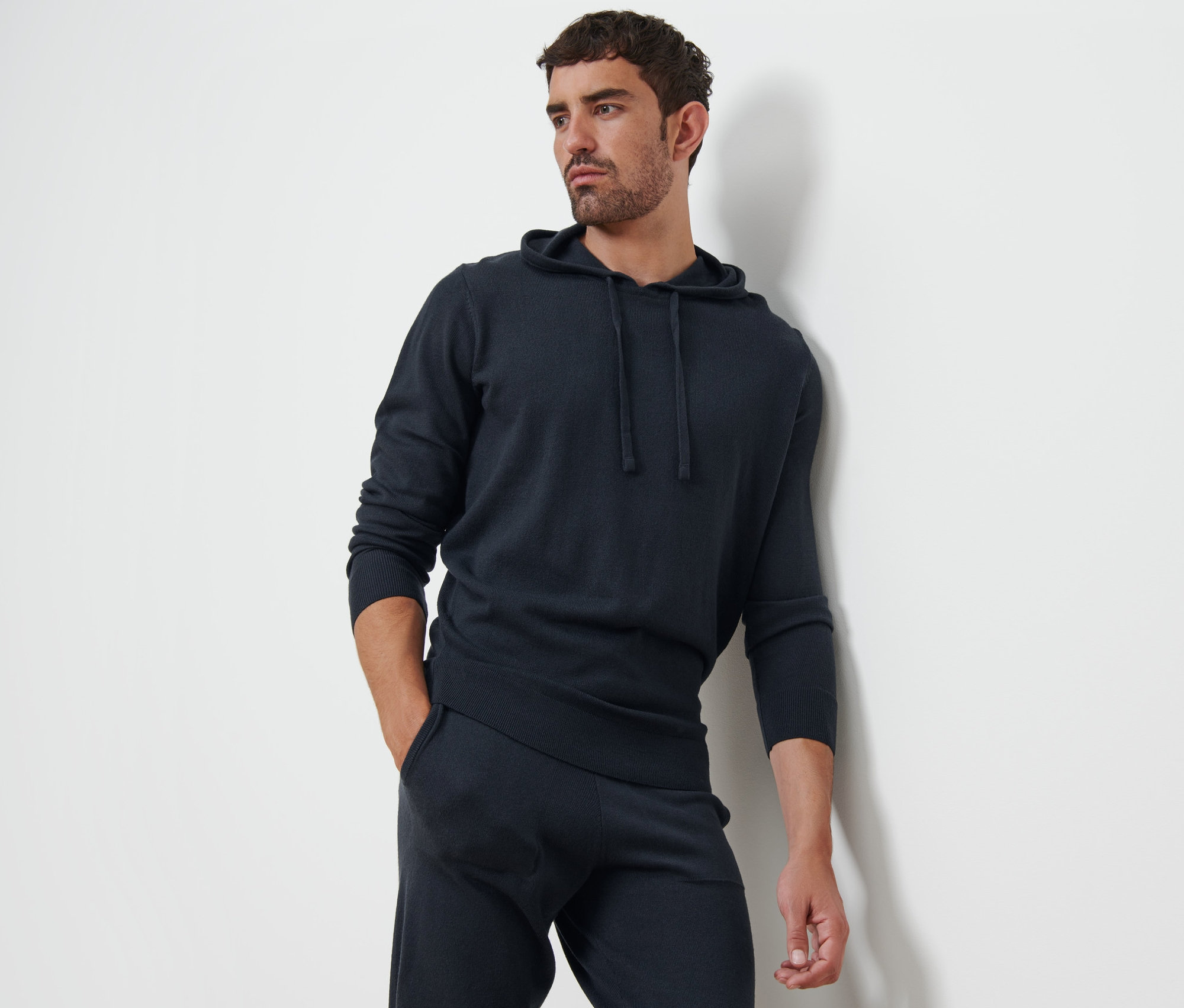Mężczyzna oparty o ścianę w bluzie dresowej z kapturem i spodniach dresowych.