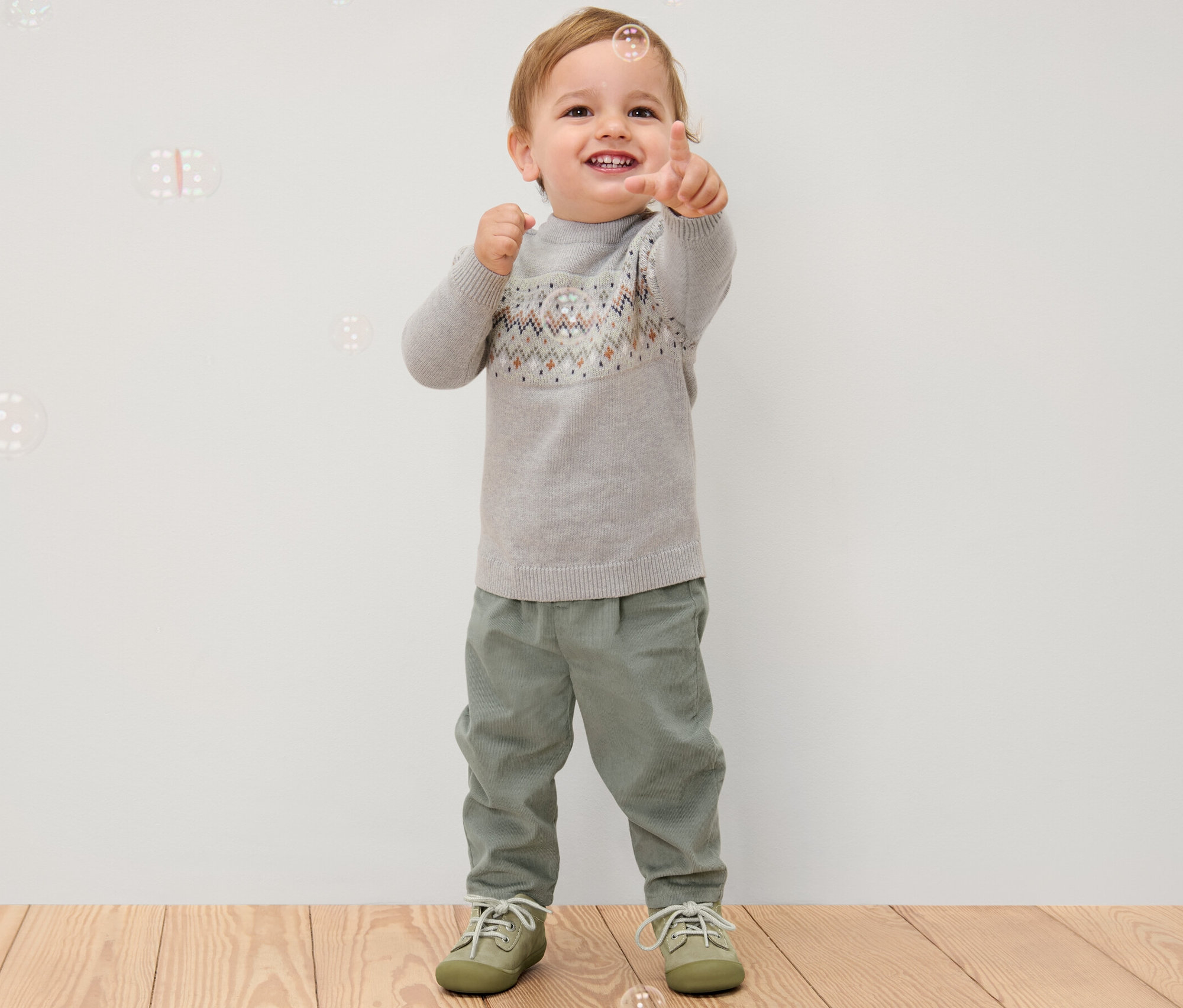Chłopiec pokazuje palcem, ubrany w szary sweter i spodnie sztruksowe na gumce.