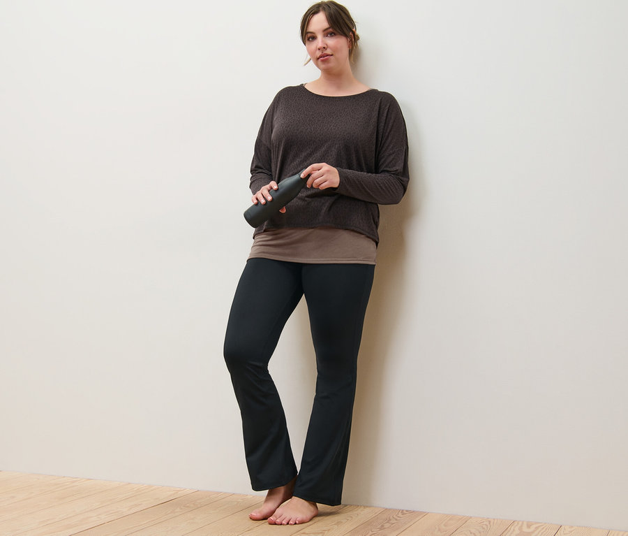 Kobieta oparta o ścianę trzyma brązową koszulkę sportową 2 w 1 i spodnie sportowe modelujące sylwetkę.