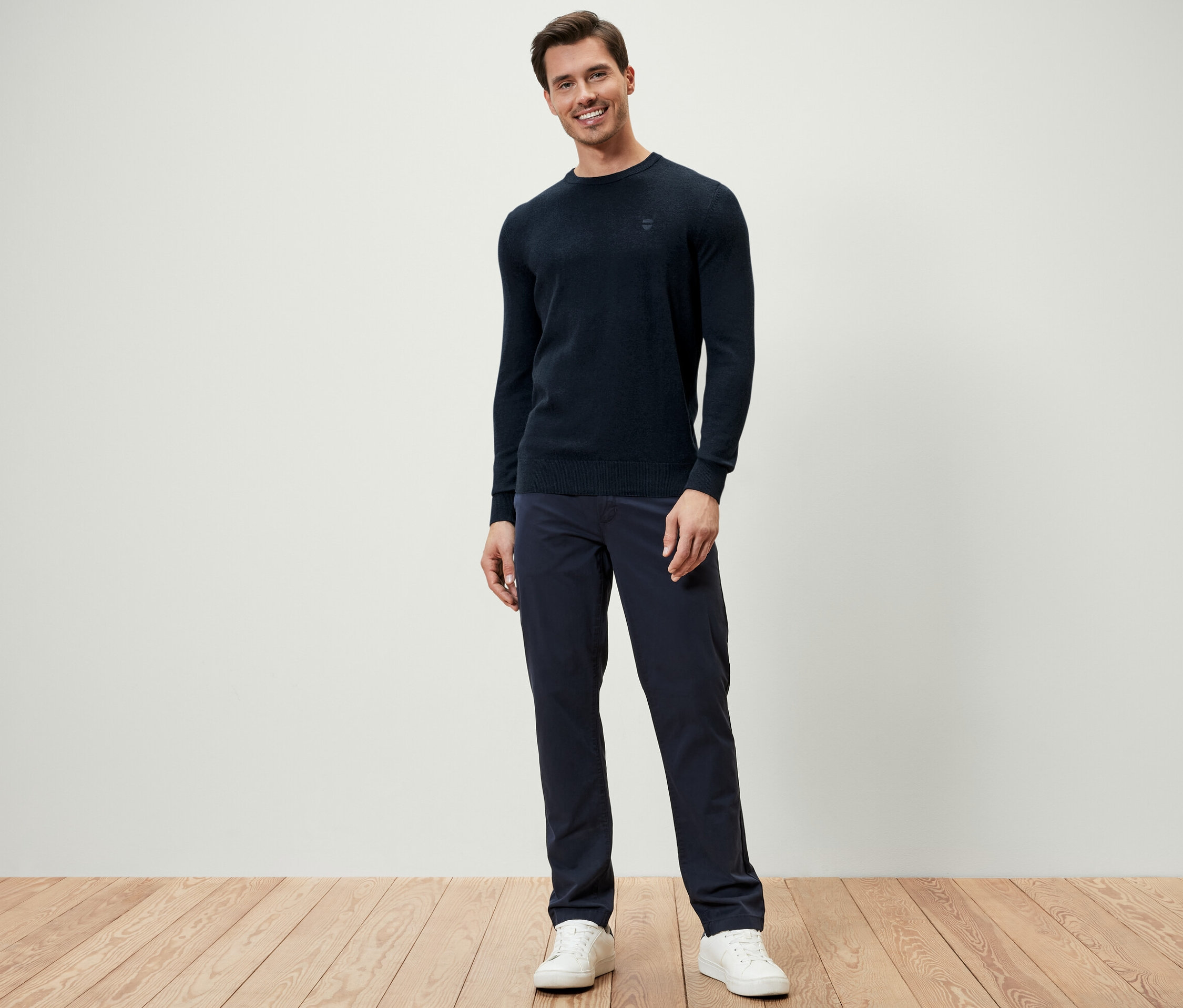 Mężczyzna stoi ubrany w granatowy sweter z okrągłym wycięciem pod szyją, granatowe spodnie i białe tenisówki.