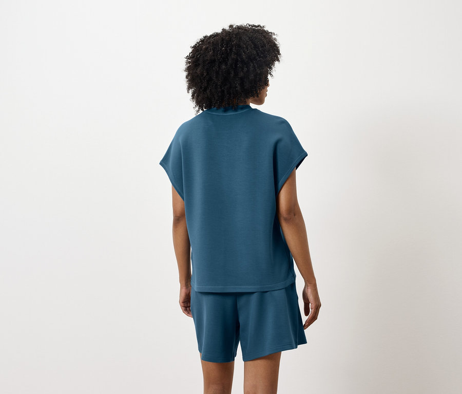 Kobieta stoi tyłem, ubrana w niebiesko-zielony top i szorty.