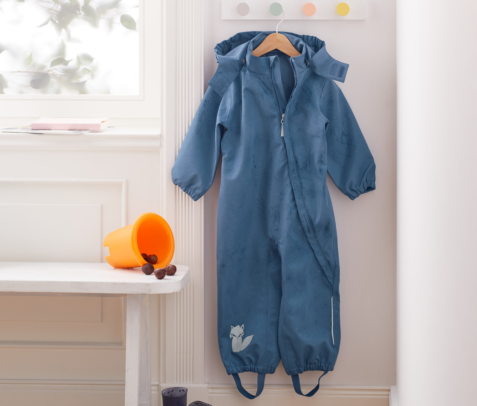 Niebieski kombinezon przeciwdeszczowy wisi na wieszaku na ścianie. Na ławce stoi pomarańczowe wiadro z kasztanami i niebieskie kalosze. W rogu stoi żółta konewka.
