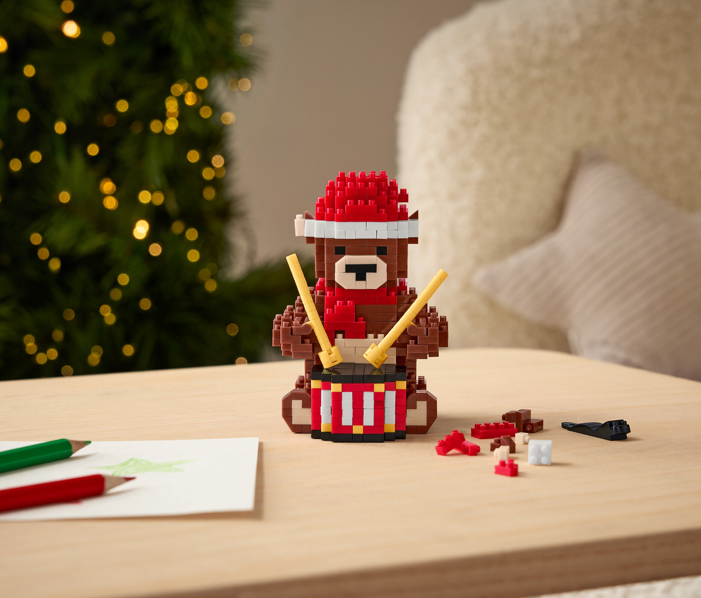 Klockowy miś z czerwoną czapką i szalikiem gra na bębnie pałeczkami na stole.