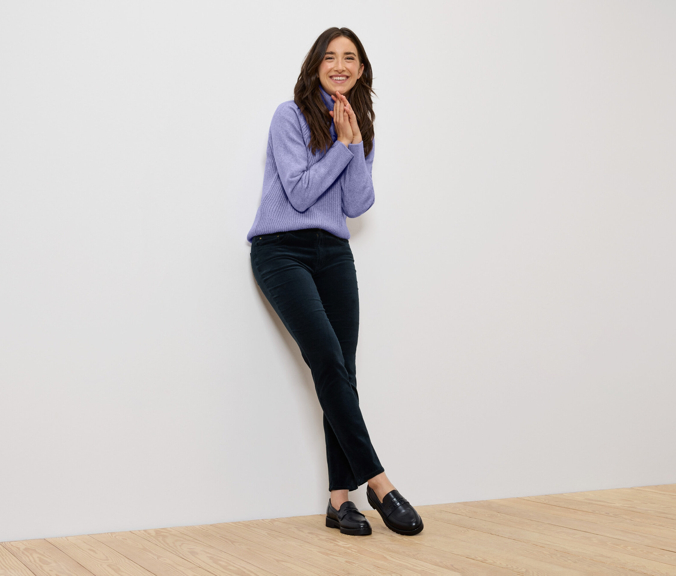 Uśmiechnięta kobieta w fioletowym swetrze i ciemnozielonych spodniach opiera się o białą ścianę.