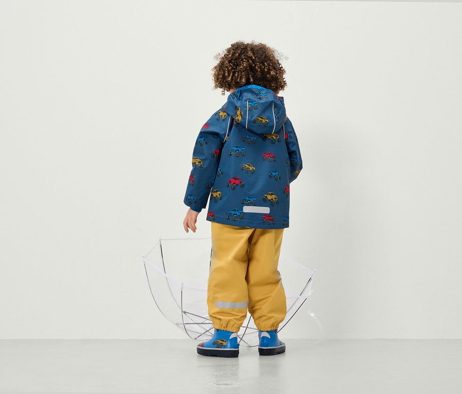 Chłopiec tyłem do kamery ma na sobie kurtkę w samochody i żółte spodnie obok przezroczystego parasola.