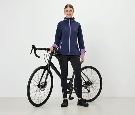 Kobieta stoi obok roweru, ma na sobie kurtkę z fioletowymi akcentami, czarne legginsy sportowe i buty.