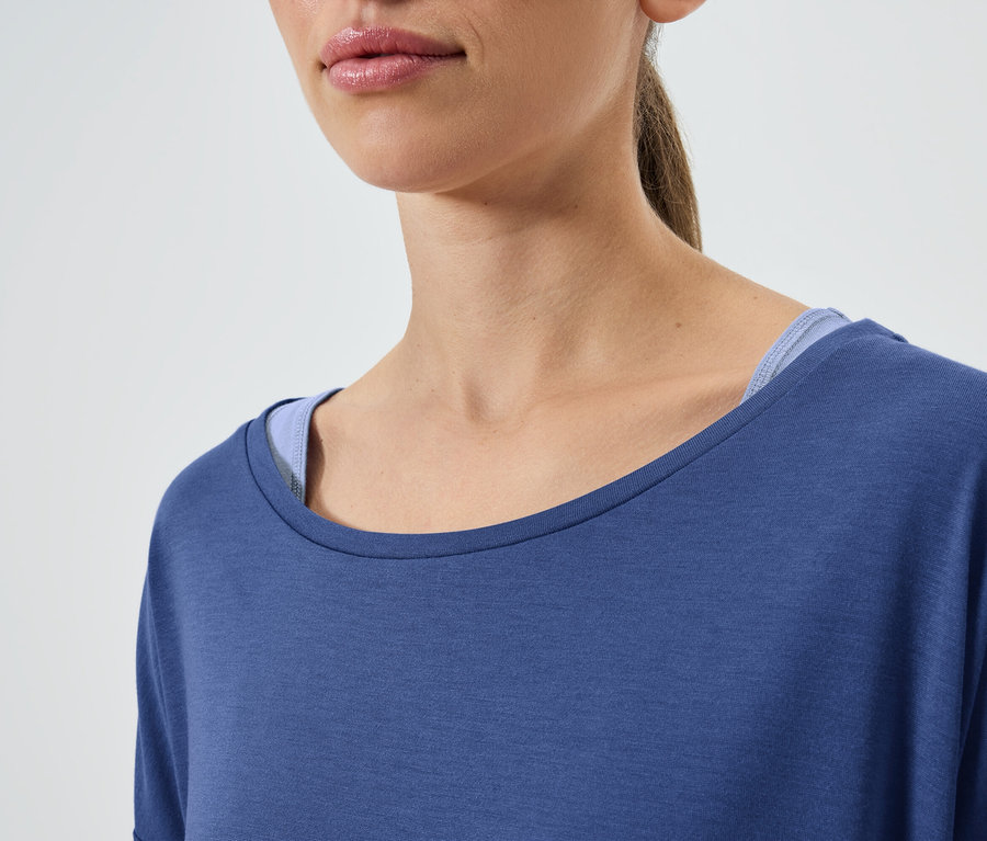 Zbliżenie na kobietę z brązowymi włosami, ubraną w niebieską koszulkę z krótkim rękawem i niebieski top na ramiączkach.