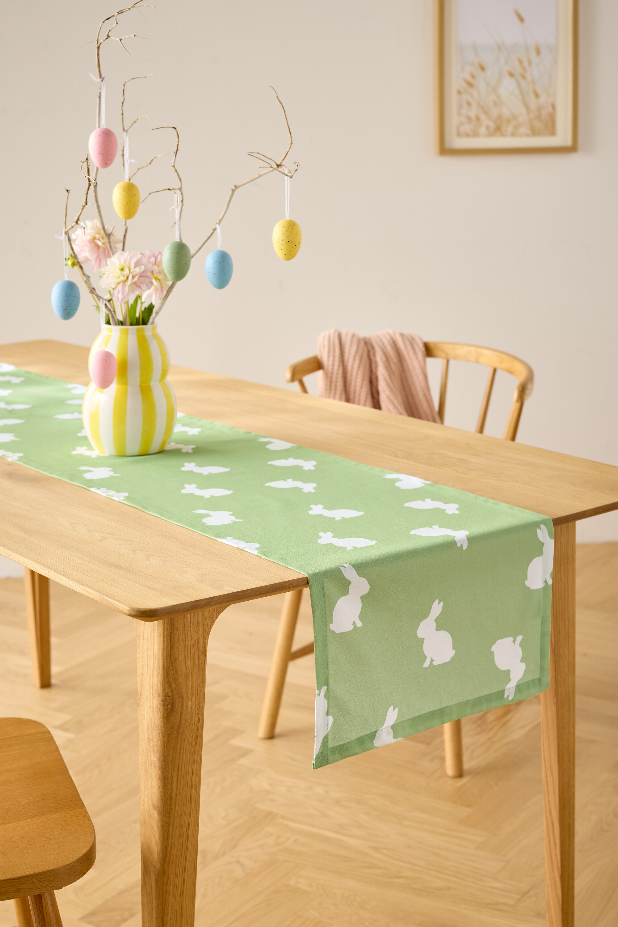 Stół jadalny z jasnozielonym bieżnikiem w białe króliczki, wazon z kwiatami i dekoracja z jajek wielkanocnych.