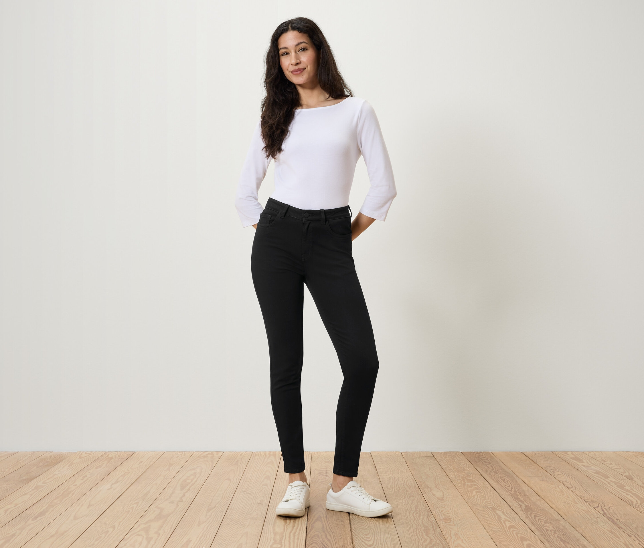 Kobieta stoi z rękami w kieszeniach czarnych dżinsów, w białej bluzce i białych tenisówkach.