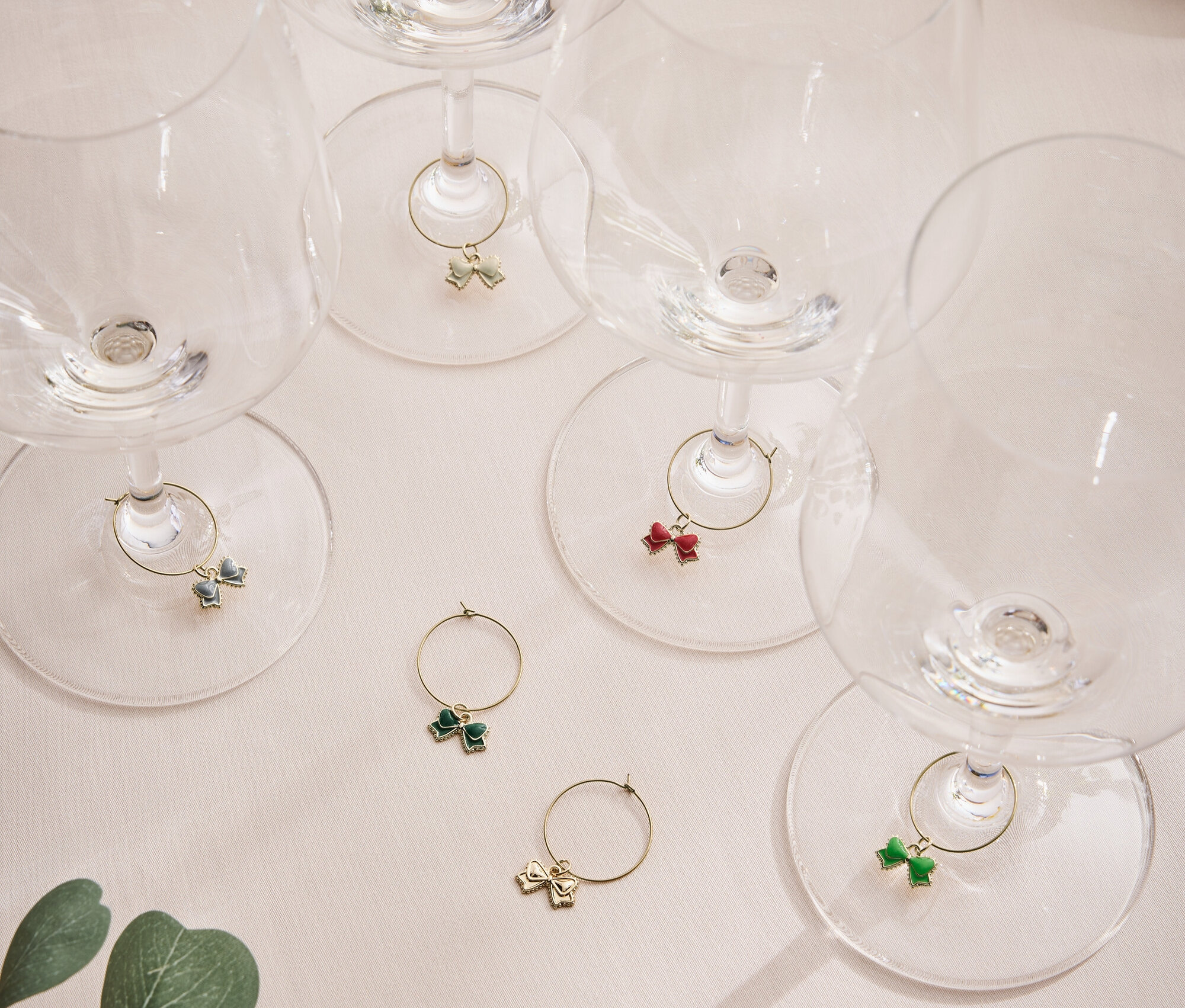 Sześć pustych kieliszków do wina z kolorowymi kokardkami na nóżkach, na jasnej powierzchni z liściem rośliny.