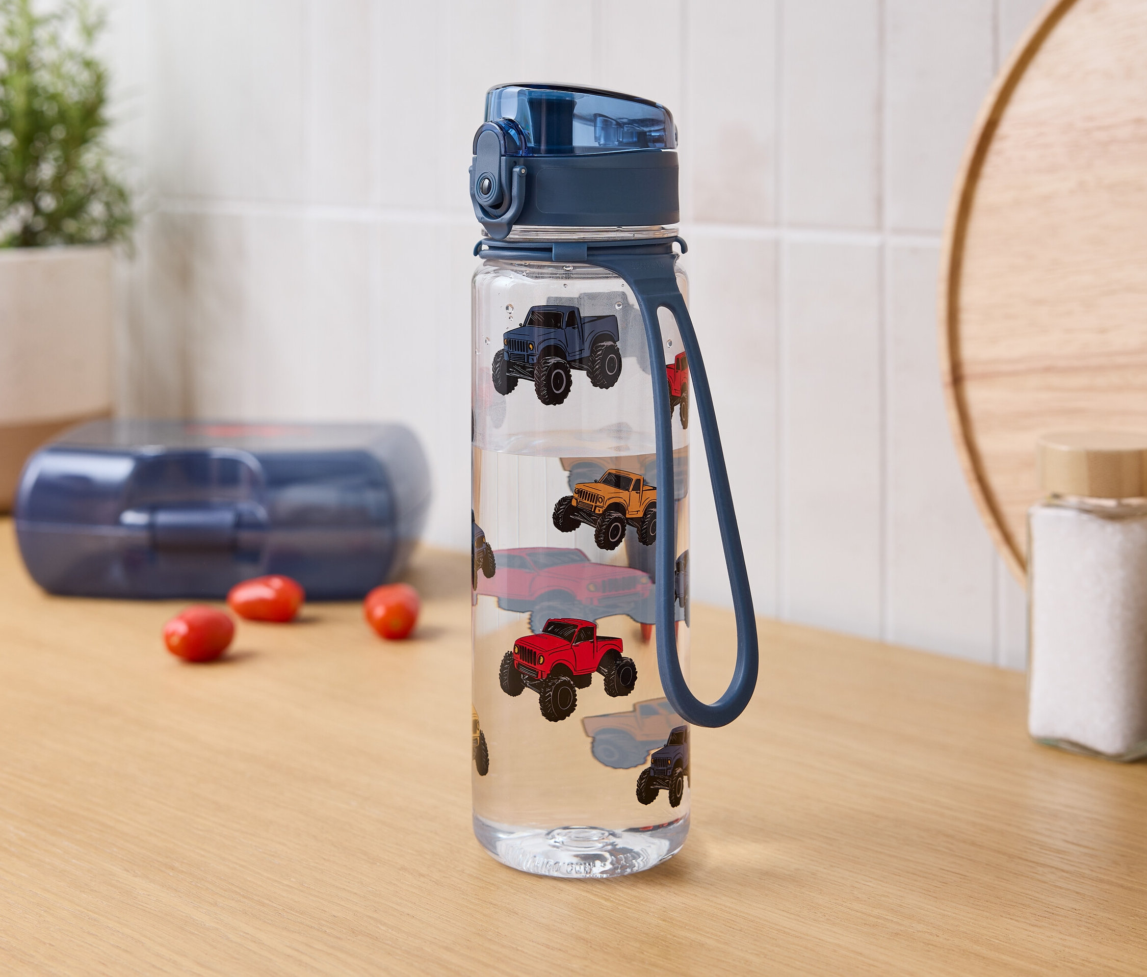 Na stole stoi butelka z wodą z nadrukiem samochodów, obok niej pudełko na lunch i przyprawy.