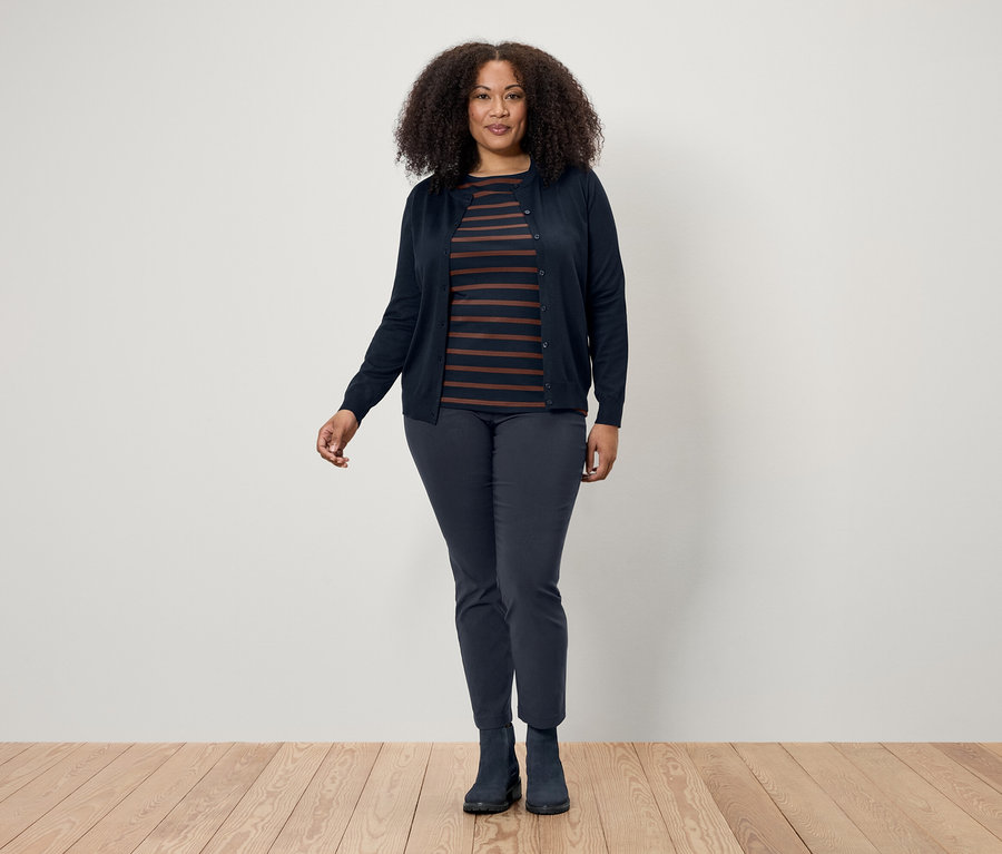 Kobieta z ciemnymi kręconymi włosami pozuje na drewnianej podłodze, ubrana w granatowy kardigan, koszulkę w paski oraz granatowe spodnie i buty.