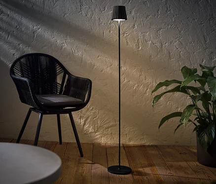 Czarna akumulatorowa lampa stojąca LED do użytku na zewnątrz stoi obok czarnego wiklinowego krzesła i rośliny doniczkowej.
