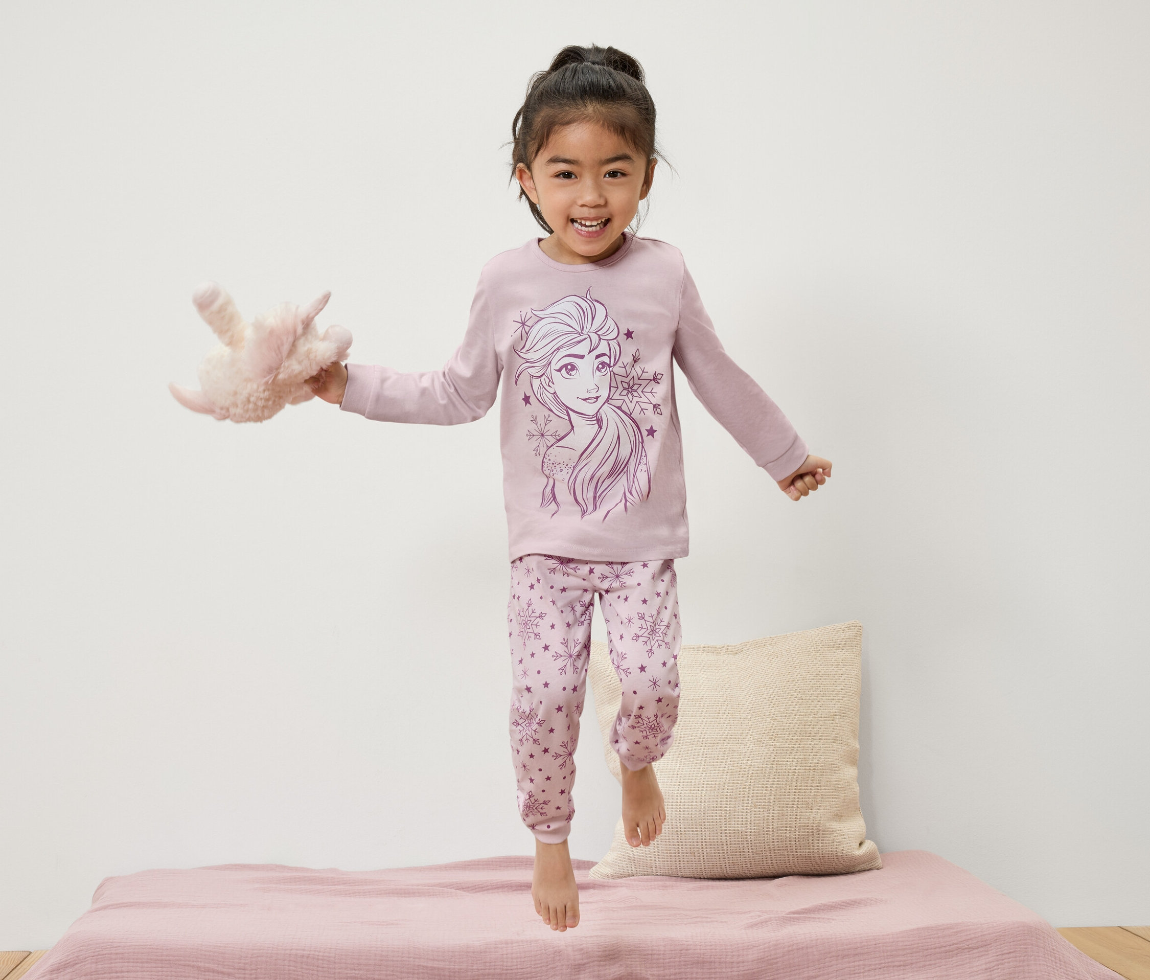 Dziewczynka w piżamie z motywem Elsy skacze na łóżku trzymając pluszową zabawkę.