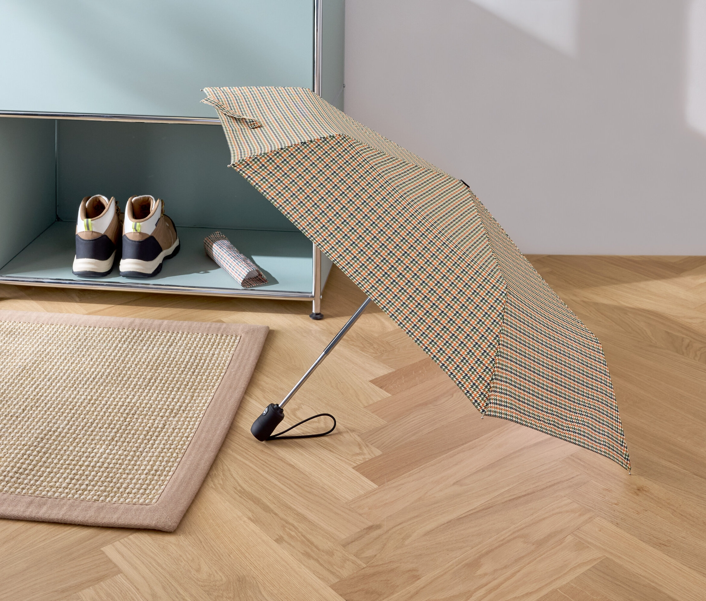 Otwarty składany parasol w kratę stoi na parkiecie. W tle widać beżowy dywan, buty i torbę na półce.