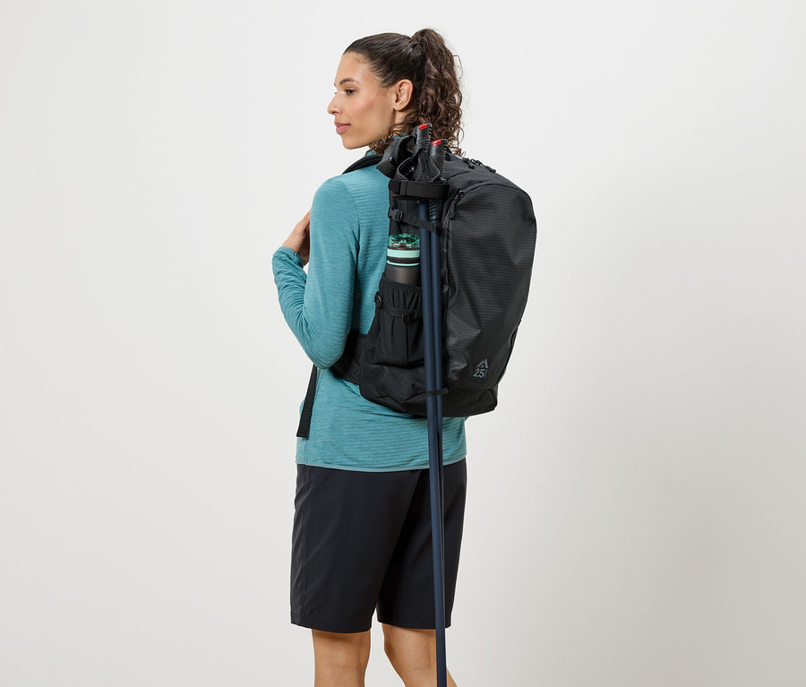 Kobieta od tyłu z czarnym plecakiem, butelką z piciem i kijkami trekkingowymi, ubrana w turkusową bluzkę i czarne szorty.