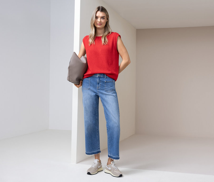 Kobieta w czerwonym topie, dżinsach i butach sportowych stoi z szarą torbą w jasnym pokoju.
