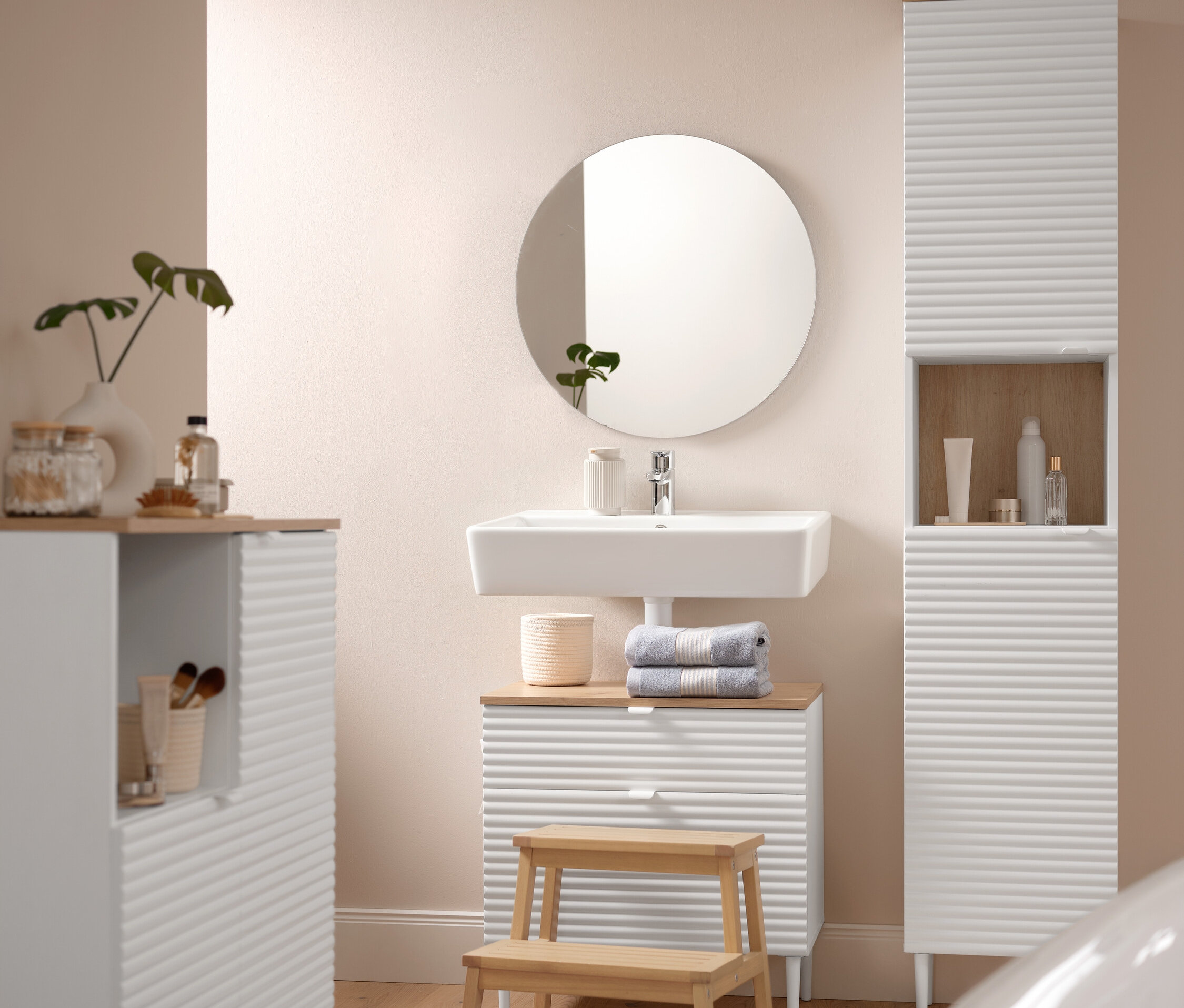 Łazienka wyposażona w szafkę wiszącą »SONARI« z drzwiami, szafką pod umywalkę »SONARI« z szufladami i komodę łazienkową »SONARI« z drzwiami. Komfortowy dywanik łazienkowy o wymiarach 60 x 100 cm leży przed stołkiem z litego drewna. Na szafce pod umywalką leżą dwa niebieskie ręczniki premium.