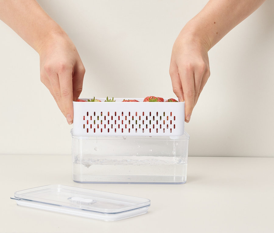 Ręce umieszczają biały pojemnik z truskawkami na przezroczystym pojemniku z wodą.