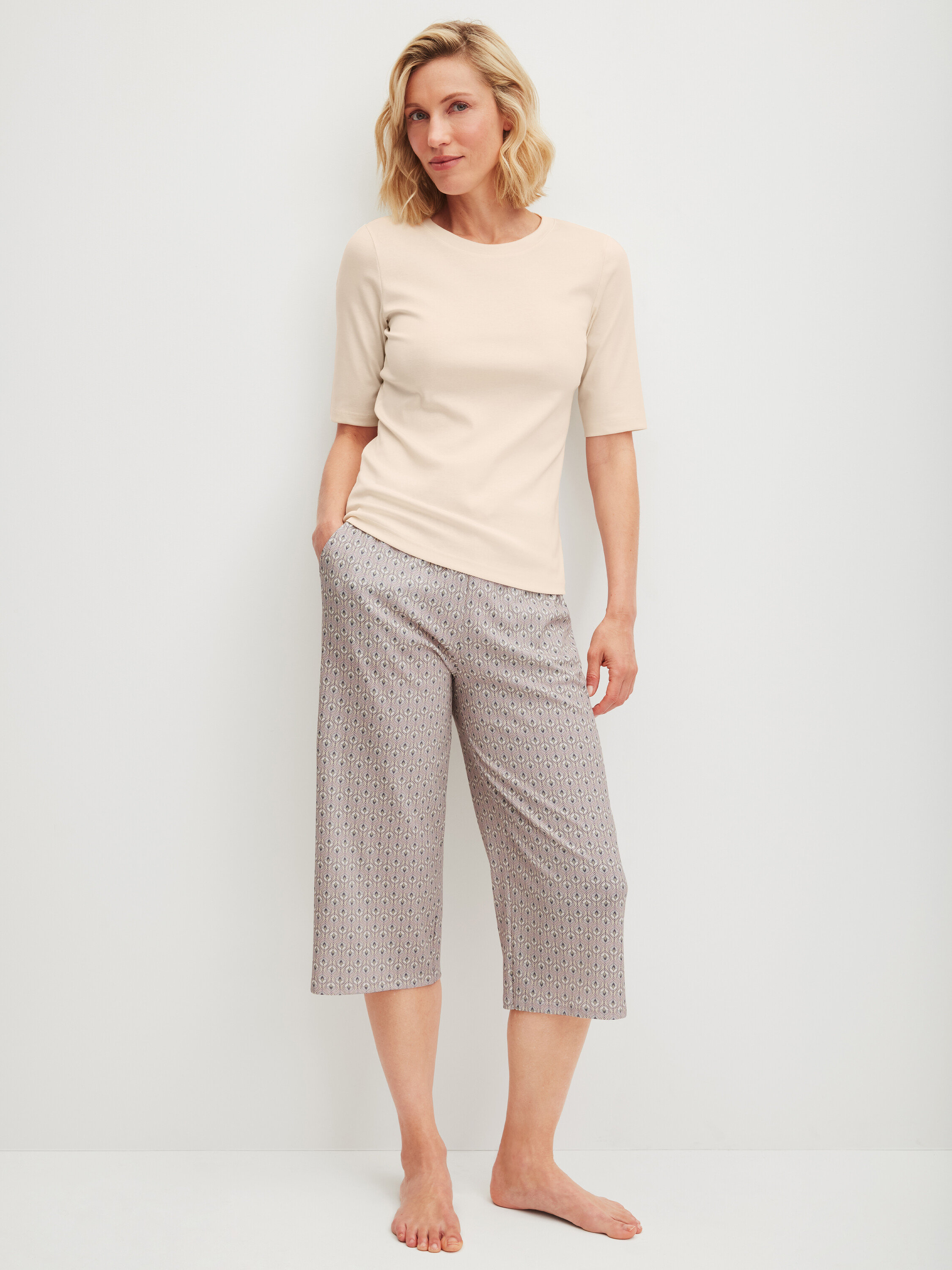 Kobieta stoi boso, ubrana w jasną koszulkę i wzorzyste spodnie 3/4.